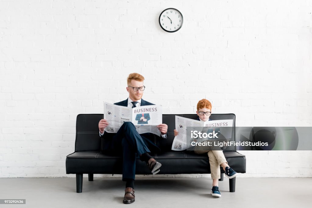 father and son businessmen wearing eyeglasses and reading newspapers on sofa Business Stock Photo