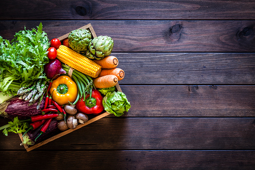 Top view of a wooden crate filled with fresh organic vegetables placed at the left of a dark wooden plank leaving useful copy space for text and/or logo at the right. Vegetables included in the composition are lettuce, tomatoes, corn, carrots, bell pepper, eggplant, asparagus, edible mushrooms, celery, artichoke and green beans. Low key DSRL studio photo taken with Canon EOS 5D Mk II and Canon EF 70-200mm f/2.8L IS II USM Telephoto Zoom Lens