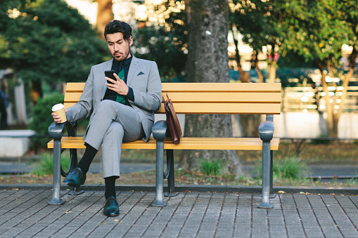 A handsome and young Japanese businessman is using a smartphone in a park in Tokyo.