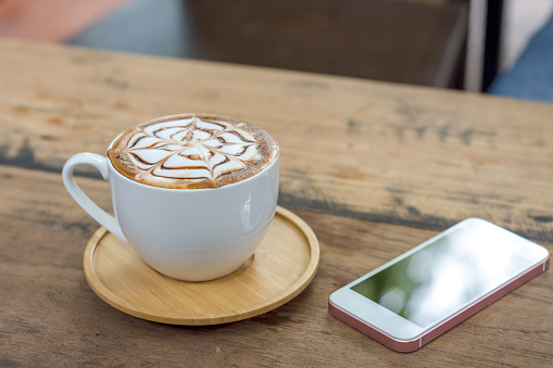 Latte art coffee cup with wooden saucer and smartphone on wood table