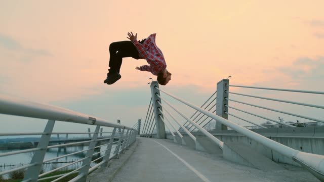 Super slow motion shot of a b-boy performing a backflip from a fence of a bridge at dusk.