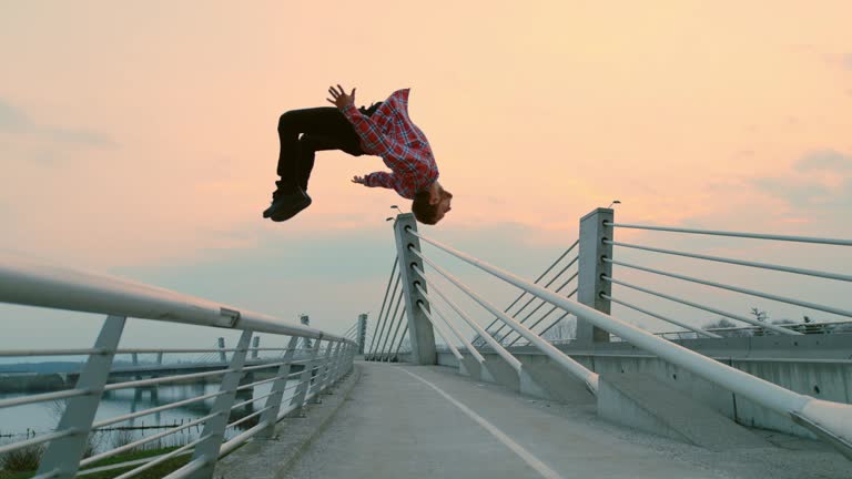 Super slow motion shot of a b-boy performing a backflip from a fence of a bridge at dusk.