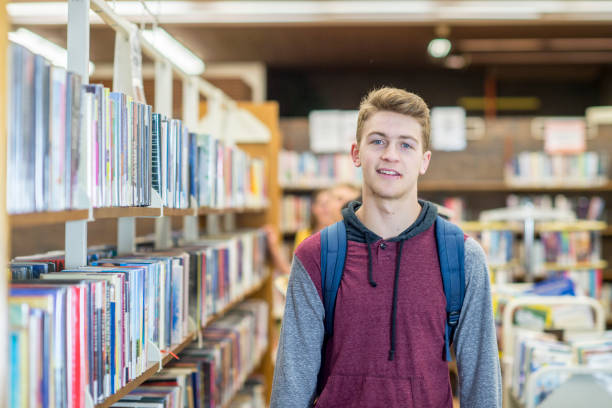 A teenage boy is indoors in his school library. He is standing and smiling for the camera while carrying his backpack.