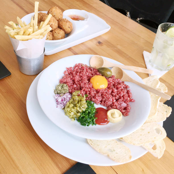 Beautiful beef tartare with egg yolk, mustard, ketchup, scallions, red onions, cornichons, capers, and bread on a white plate in a Sao Paulo, Brasil restaurant.  On table with croquettes, fries, and caipirinha.