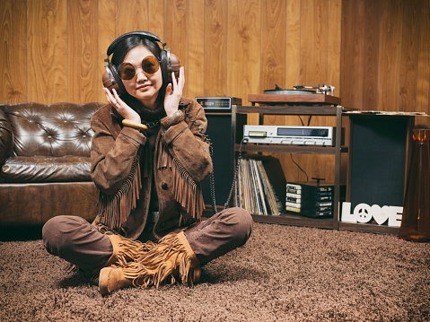 Above view of young woman in jeans and shirt looking at record player with vynil disk while listening to music in headphones on the floor