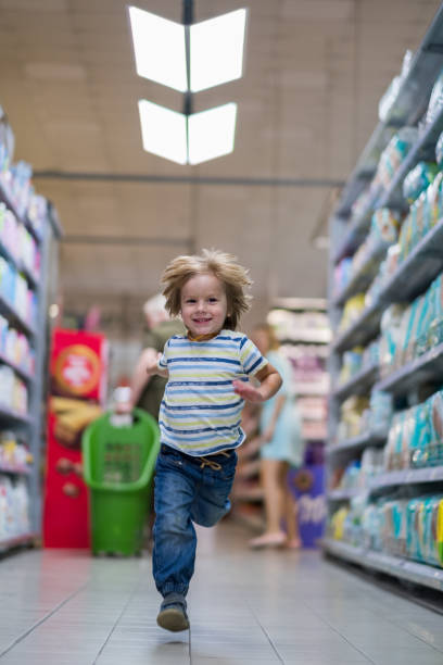 A sleepy and cute Eurasian one year old boy looks directly at the camera while sitting in a shopping cart while shopping with his parents in the clothing section of a superstore.