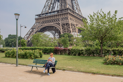 Paris / France - May 15, 2018: A young woman enjoys a quiet moment sitting on a bench in front of the iconic French landmark, the Eiffel Tower.