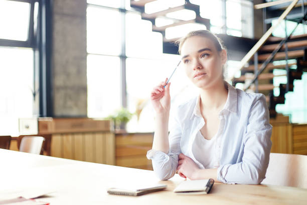 istock Young woman in white blouse contemplating on assignment while sitting over notepad at desk with pen in her hand 973488964