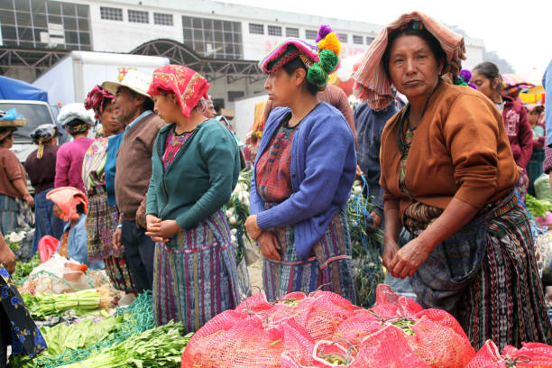 Guatemala, La Antigua - July 20, 2023: Local women in colorful traditional clothing sit in park of Plaza Mayor, central square, with child and other people around. Eating while selling toys