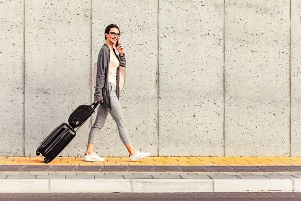 istock Young woman using her smart phone walking beside the concrete wall and pulling a small wheeled luggage with a briefcase on it 973515624