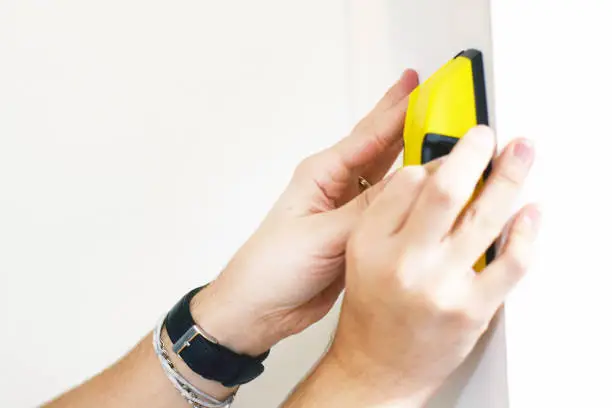 Photo of Man using stud finder to search apartment wall for studs and live wires