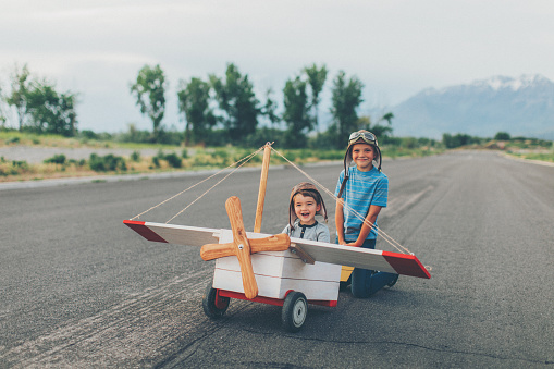 Two young boys dressed in flight caps and goggles get ready to fly a vintage toy airplane. One boy is sitting in the plane as pilot while the other is getting ready to push the plane. Seeing sights from the sky has never been better. Both boys are smiling and looking at the camera. Image taken in Utah, USA.