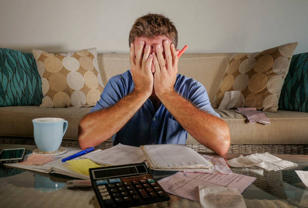 worried and stressed man at home calculating month tax expenses with calculator accounting payments doing bank paperwork in stress desperate suffering financial problem covering face stock photo