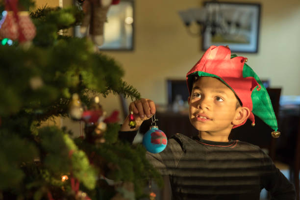 High quality stock photo of a young boy decorating the Christmas tree in the morning with natural light and a homemade Santa Claus hat.