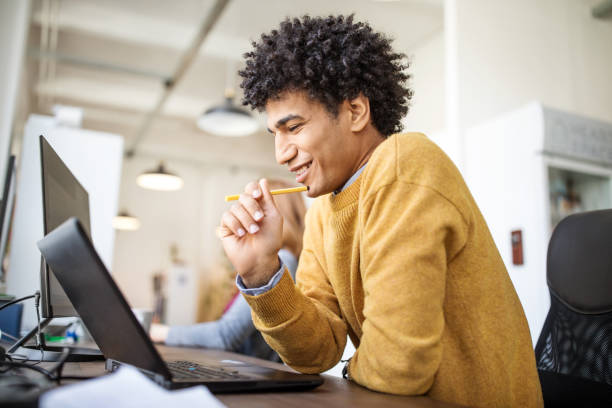 Smiling businessman working at his desk Side view if happy young businessman sitting at his desk and working on laptop computer. Egyptian male professional looking at his laptop and smiling in office. egyptian ethnicity stock pictures, royalty-free photos & images