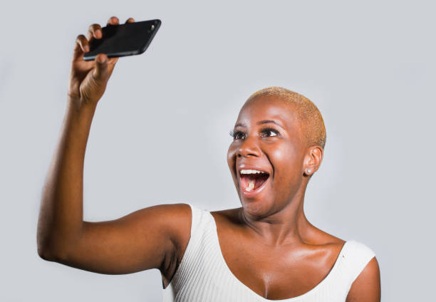 孤立した背景上若い美しく、幸せな黒アフロ ・ アメリカ人女性の携帯電話で selfie の写真肖像画を撮影または録音セルフ ポートレート ビデオ陽気なポーズに興奮して笑顔で楽しんでくださ� - shaved head ストックフォトと画像