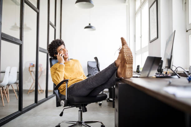 Relaxed businessman talking on phone in office Smiling businessman talking on mobile phone while sitting on chair and feet up on table. Relaxed male professional taking a break from work in office talking on phone. feet up stock pictures, royalty-free photos & images