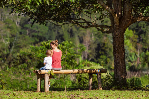 mère avec fils s’asseoir sur le banc sous l’arbre - tree grass family human relationship family photos et images de collection