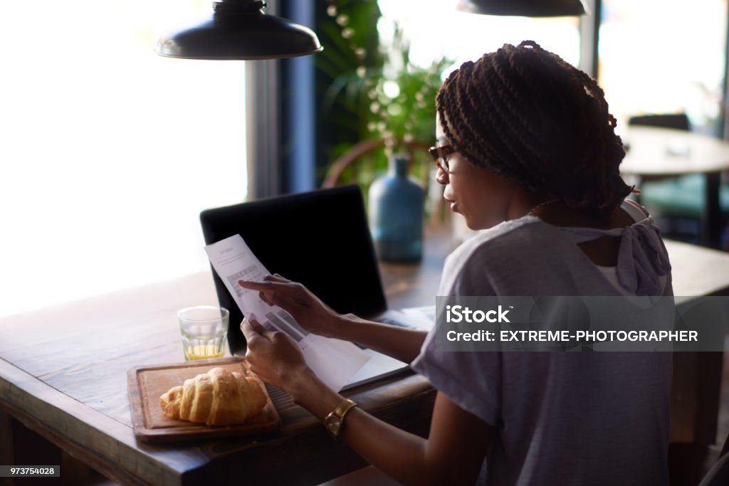 Woman with papers Young black woman with eyeglasses holding paper documents. Laptop computer and croissant are on the table. Adult Stock Photo