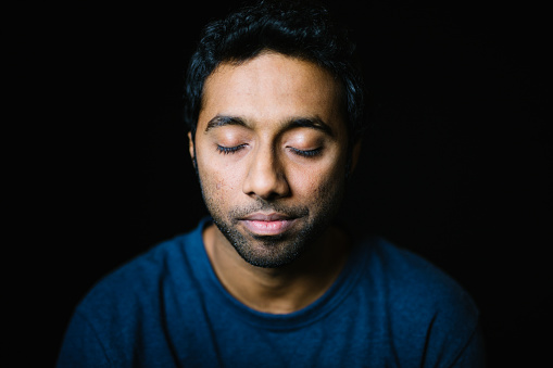 Close-up of young man with eyes closed. Portrait of thoughtful male is wearing blue T-shirt. He is on black background.