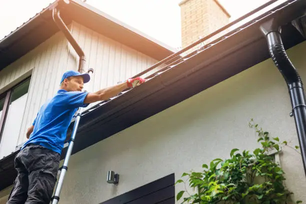 Photo of man on ladder cleaning house gutter from leaves and dirt