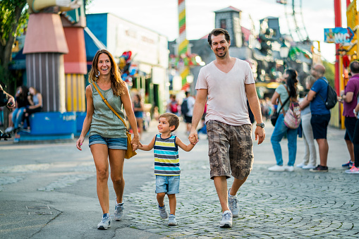 little son with smiling mother and father walking hand in hand together through amusement park in summer