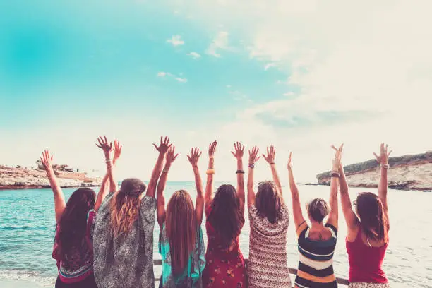 group of women enjoying and celebrate summer vacation all together saying hallo to the ocean and the nature. young people leisure activity, all hands up and vintage colors style. fashion dresses view from backside