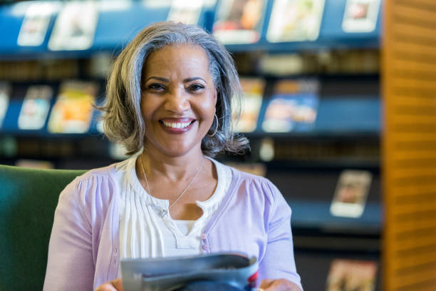 Beautiful senior woman reading magazine in library Beautiful senior African American woman reads a magazine while in the library. She is smiling cheerfully at the camera. librarian stock pictures, royalty-free photos & images