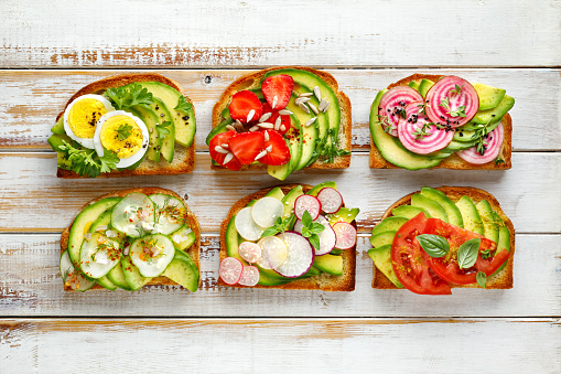 Avocado sandwiches, toasts with various vegetarian toppings on a white wooden table, top view