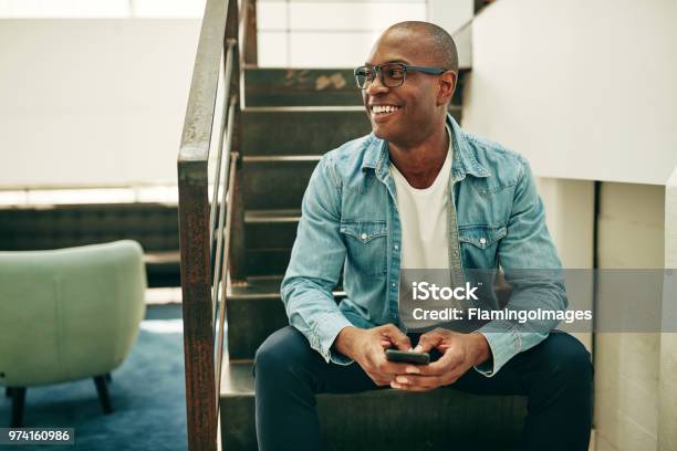 Uomo Daffari Africano Sorridente Seduto Sulle Scale Dellufficio Con In Mano Un Cellulare - Fotografie stock e altre immagini di Uomini