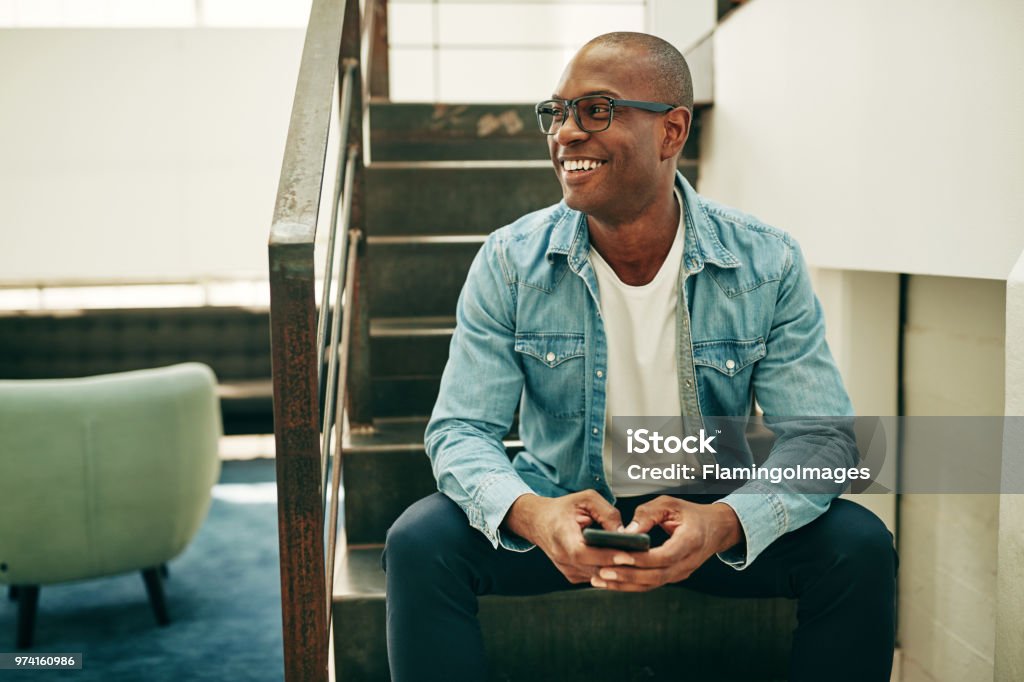 Smiling African businessman sitting on office stairs holding a cellphone Smiling young African businessman wearing glasses and reading text messages on a cellphone while sitting on stairs in an office Men Stock Photo