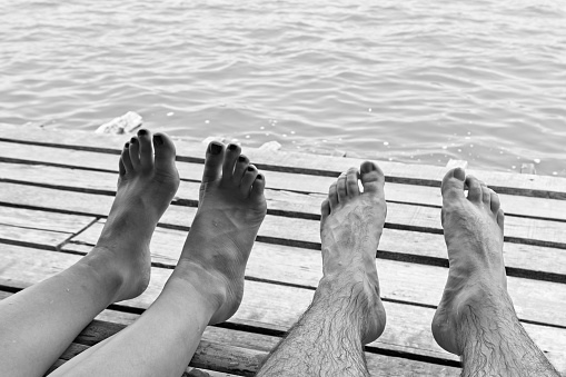 A black and white close up shot of a Caucasian couple's feet while sitting on a jetty. This image can be used to represent love between two people.