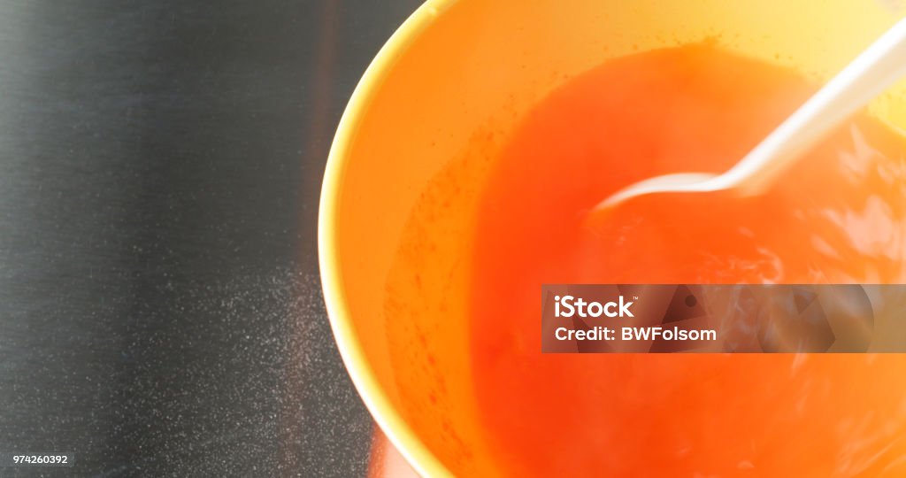 Stirring orange gelatin mix into a bowl of hot water with a white plastic spoon Close view of stirring orange gelatin mix into a bowl of hot water with a white plastic spoon on a stainless steel counter top. Bowl Stock Photo