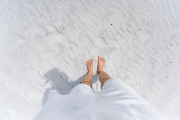 Young woman looking down pov point of view perspective on bare feet standing in white sand in Fort Walton Beach, Florida, in white dress, flat top view with shadow