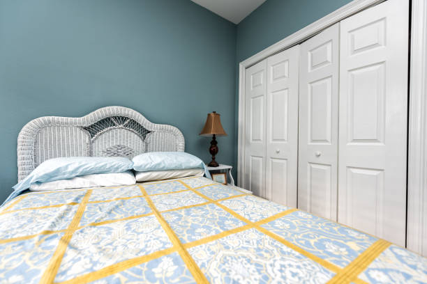 istock New clean bed comforter with headboard, table, lamp, vintage beach theme decorative blue green pillows in bedroom in staging model home, house or apartment by closet 974310766