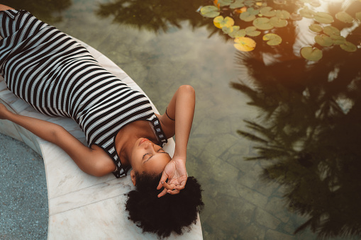 Tired beautiful pregnant African-American girl with curly afro hair is resting and laying on the marble bench near the pond with waterlilies on a sunny summer morning, palms silhouettes in the water