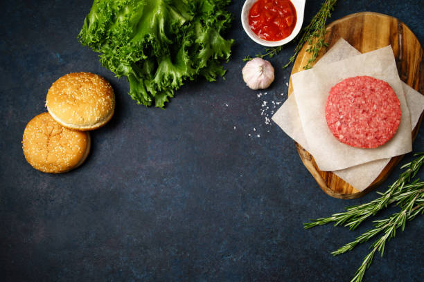 istock Cooking burger: classical american burger ingredients on dark blue background with copy space. Raw beef patties, vegetables, buns, lettuce. 974363834