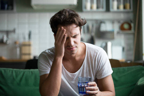 Young man suffering from headache, migraine or hangover at home Young man suffering from strong headache or migraine sitting with glass of water in the kitchen, millennial guy feeling intoxication and pain touching aching head, morning after hangover concept headache photos stock pictures, royalty-free photos & images