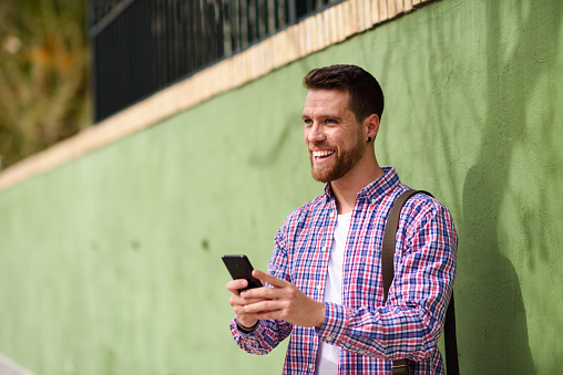 Young man laughing with his smart phone in urban background. Guy wearing casual clothes. Lifestyle concept.