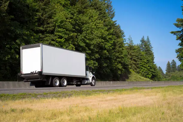 Photo of Big rig semi truck with long box trailer running on green road with trees and blue sky