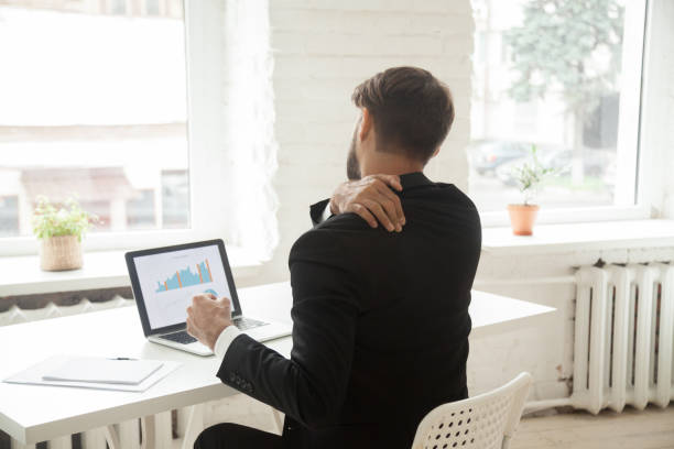 Tired businessman stretching at workplace exhausted after work Exhausted businessman stretching at workplace in office, tired after long working hours. Male entrepreneur massaging back suffering from strain, sedentary work and uncomfortable sitting. Back view office back pain stock pictures, royalty-free photos & images