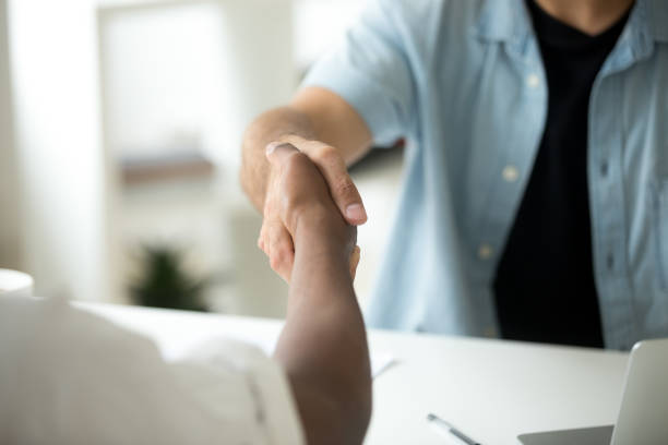 Close up of multiracial handshake over office desk Close up of multiracial handshake at office desk. Caucasian worker shaking hand of black colleague, welcoming at new workplace, greeting, being promoted. Cooperation, support, making agreement concept casual job interview stock pictures, royalty-free photos & images