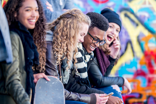 A group of teenagers are sitting in front of a wall covered in graffiti. They are wearing stylish clothes. Everyone is enjoying spending time together.
