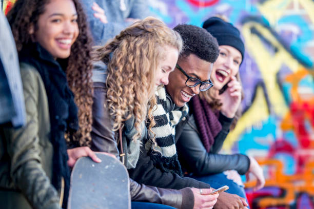 Teens In Urban Environment A group of teenagers are sitting in front of a wall covered in graffiti. They are wearing stylish clothes. Everyone is enjoying spending time together. mural photos stock pictures, royalty-free photos & images