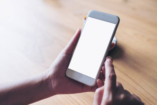 istock Mockup image of hands holding white mobile phone with blank desktop screen on wooden table 974676354