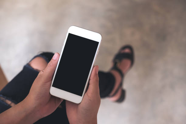 istock Top view mockup image of a woman's hands holding white mobile phone with blank black screen on thigh 974676410