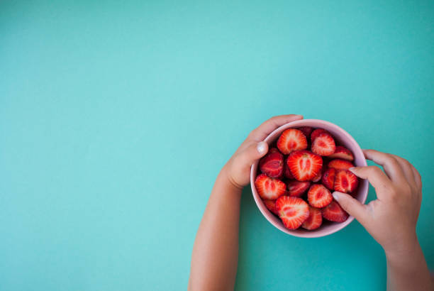 Child's hands holding a pink bowl full of beautiful fresh juicy strawberries on pastel blue background with text space.