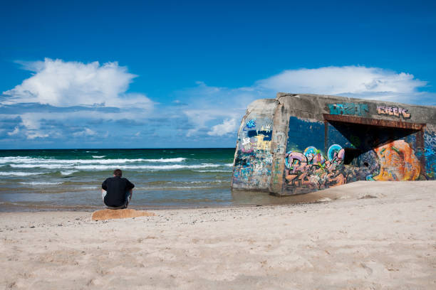 This image was made in Soulac sur Mer beach, in Nouvelle Aquitaine, in  France. It was made in July 20, 2014. There is a Bunker on the beach, in the sea. It's decorated with painting very colored. On the left, a man is sitting down, on the beach. He is looking the sea.