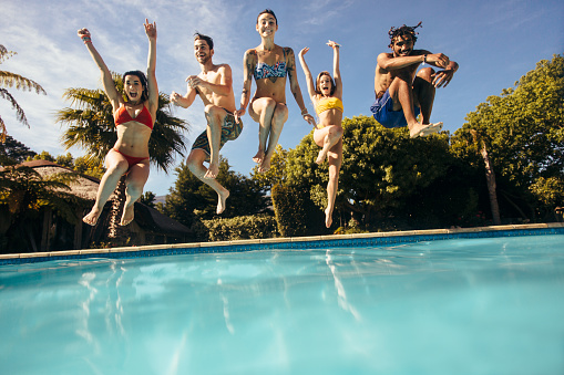 Happy young friends jumping into outdoor swimming pool and having fun. Group of men and women jumping into a holiday resort pool.