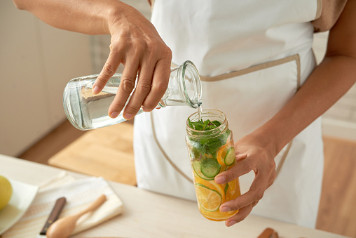 Close-up shot of unrecognizable woman wearing apron preparing citrus and mint infused water while standing at modern kitchen illuminated with sunbeams